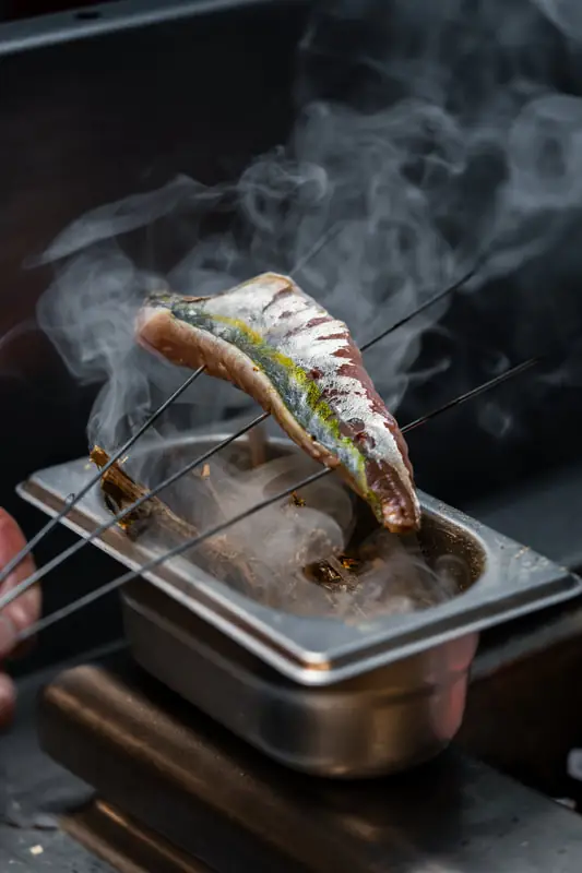 A mackerel fillet held on metal skewers over a smoking metal tray of burning embers