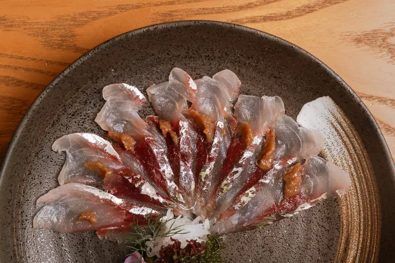 Thin slices of white fish sashimi fanned in a circle over a red sauce on a dark speckled plate