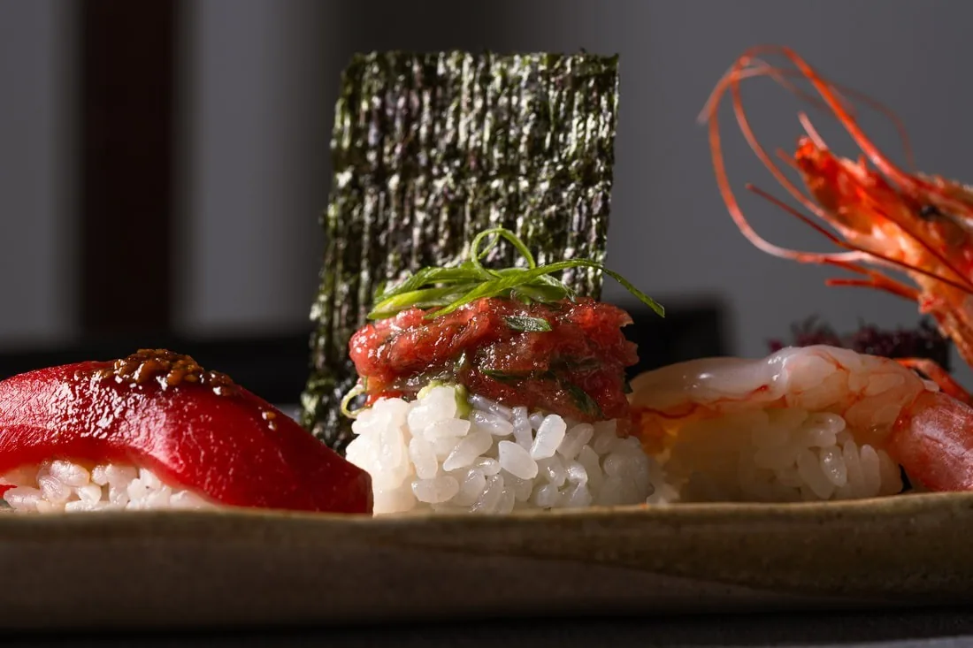 Three pieces of nigiri, including tuna, a tartare topped one with a strip of seaweed, and a prawn, on a ceramic plate