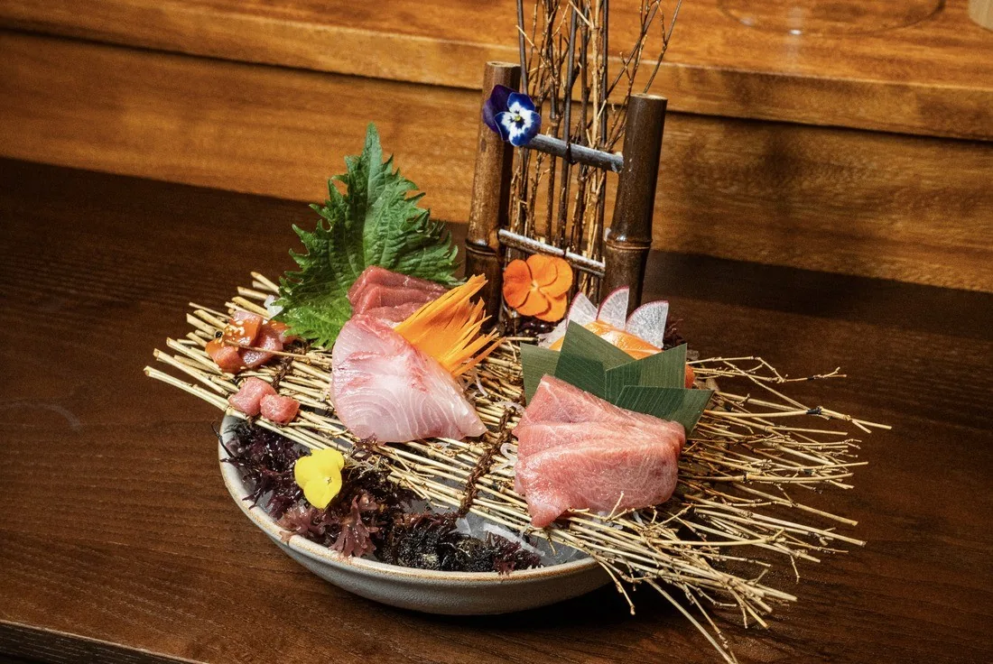 A bowl of sashimi with pink and white fish, herbs and dried grass garnish on a wooden table