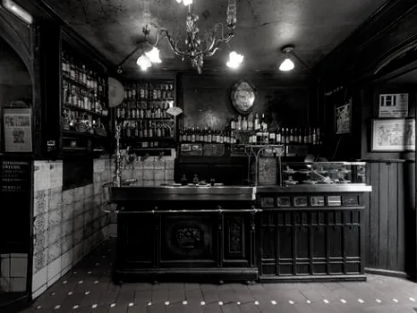 The tavern's dark carved wooden bar counter in black and white, tiled dado behind, shelves of bottles, a chandelier overhead and a tiled floor