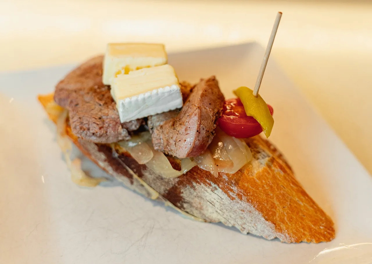 A slice of bread topped with meat, cheese and a pickled pepper on a skewer