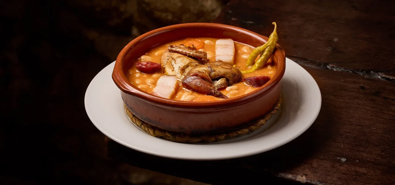 A clay bowl of stew on a white plate on a dark wooden table