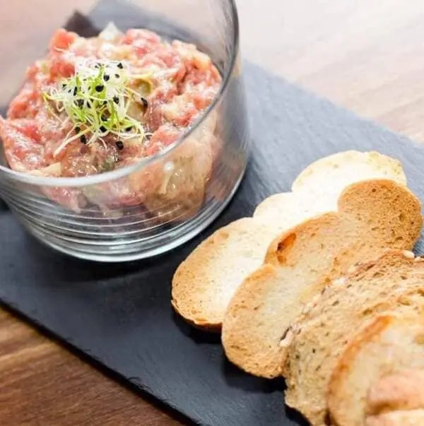 Steak tartare topped with microgreens and black sesame in a glass tumbler on a slate board, with sliced toasts alongside