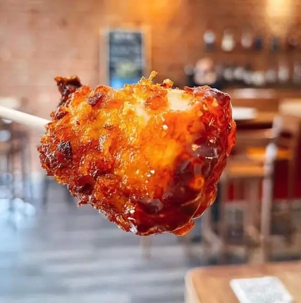 Close-up of a glazed fried croquette on a skewer held up against a blurred exposed-brick bar interior