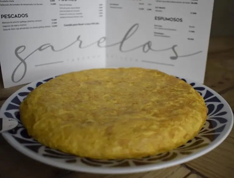 Thick golden Spanish potato omelette (tortilla) on a blue-and-white patterned plate, with a printed Garelos menu propped behind it on a wooden table