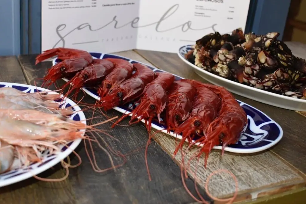 Wooden table with a plate of large red carabinero prawns, a plate of raw pale prawns, and a plate of goose barnacles (percebes), with a Garelos menu behind