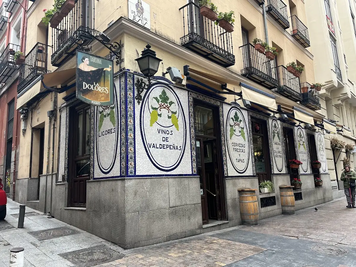 Corner exterior of Taberna La Dolores in Madrid, ornate blue-and-white tiled facade with hanging sign and oval mosaics reading Vinos de Valdepenas and Cervezas Frescas y Tapas, wine barrels by the door