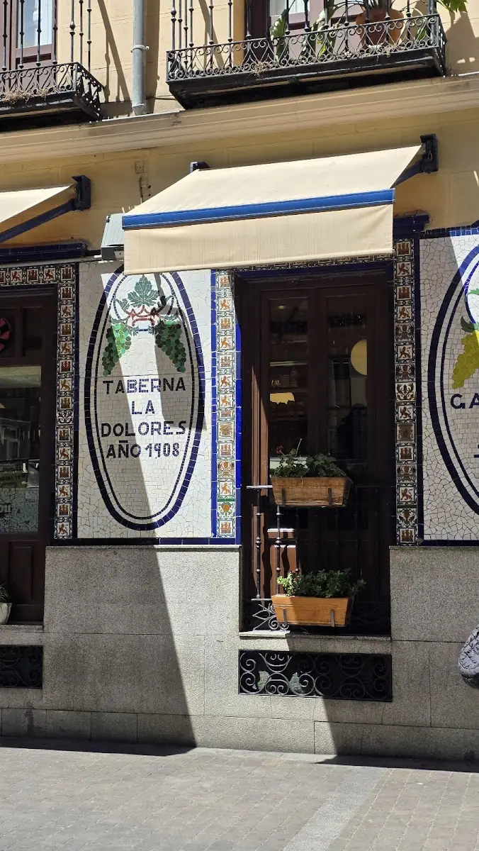 Tiled tavern facade with a blue-and-white mosaic panel reading Taberna La Dolores Ano 1908, a cream awning and window boxes of plants