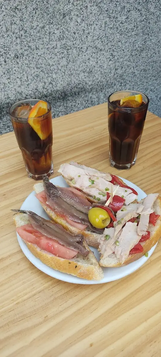 Plate of pintxos on baguette slices topped with anchovies, tuna, tomato and a skewered olive, with two tall glasses of vermouth garnished with orange on a wooden table
