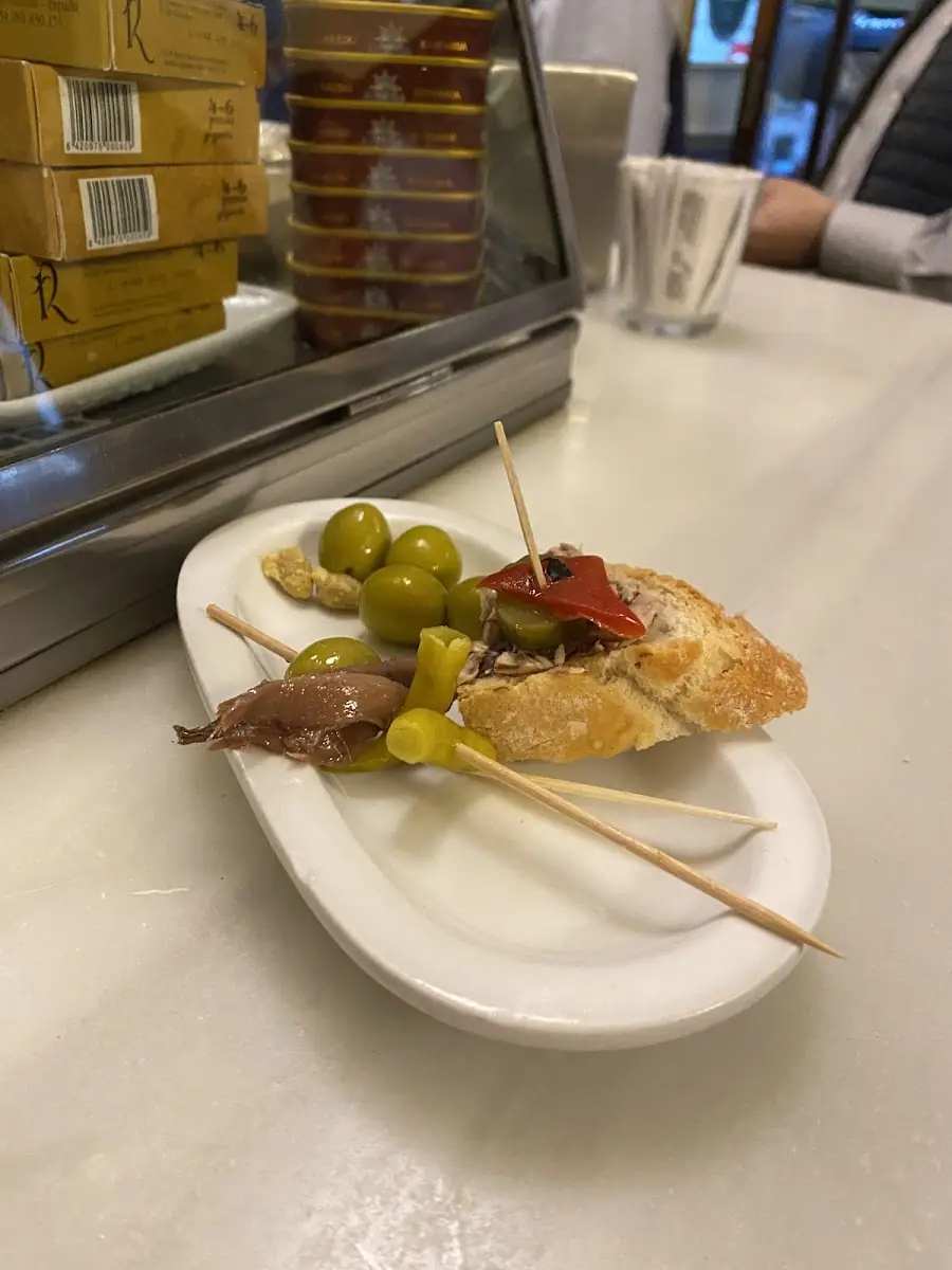 Small oval plate with an anchovy-and-olive skewer, green olives and a tuna-and-piquillo-pepper pintxo on bread at a marble bar top