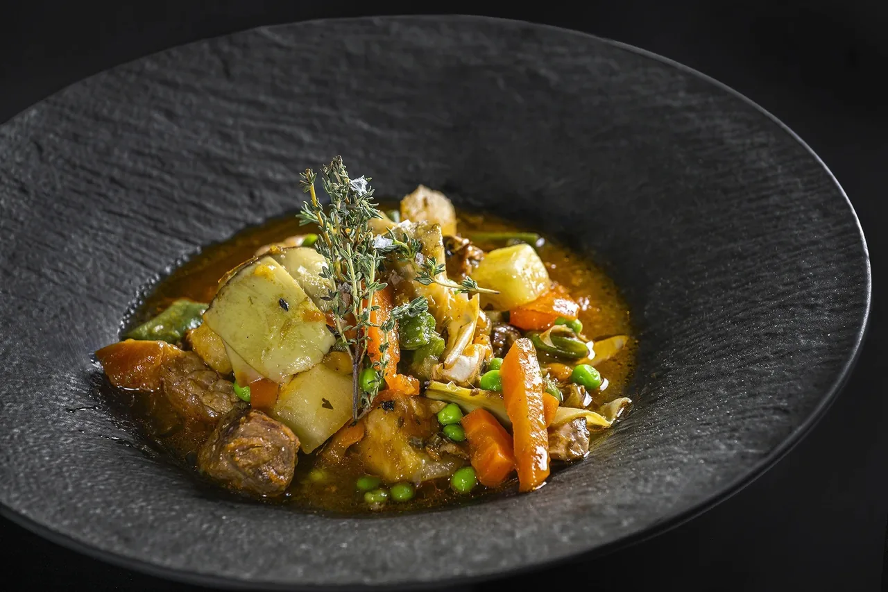 A dark shallow bowl of vegetable stew with potatoes and peas