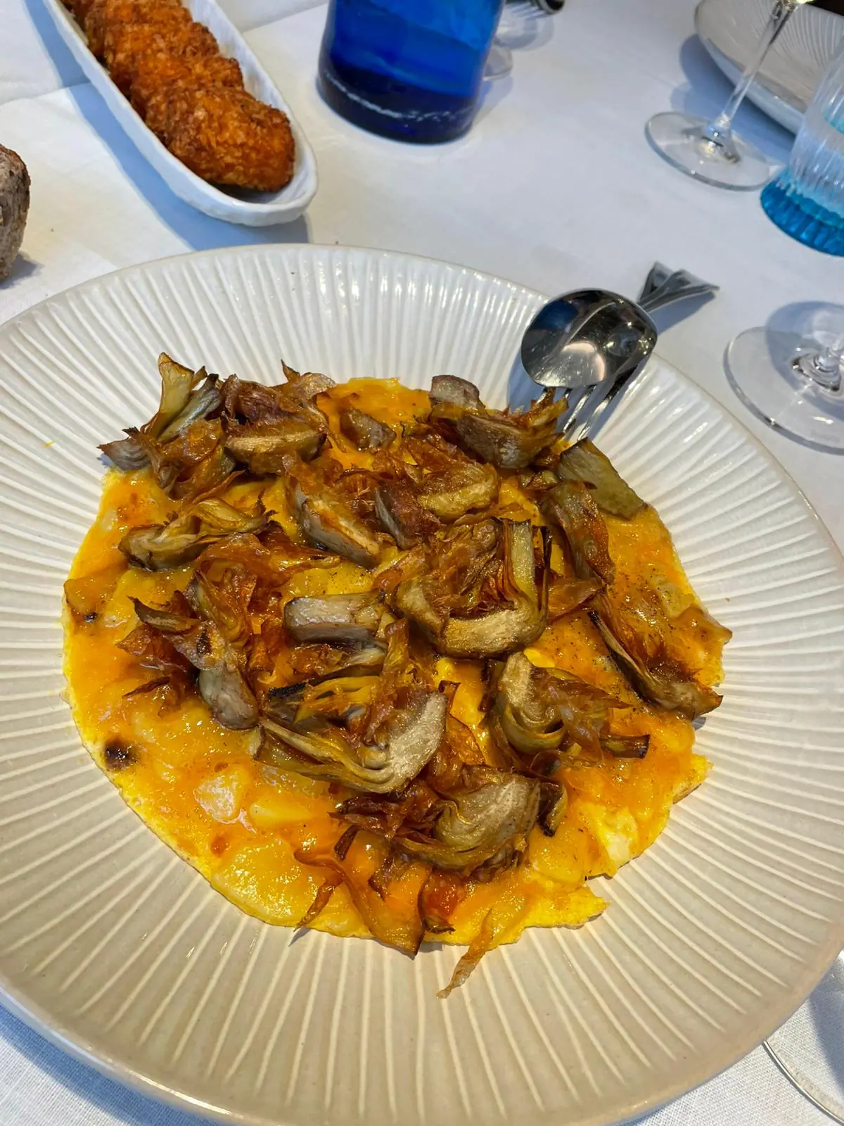 Baked egg dish topped with fried artichoke pieces on a plate, croquettes visible in background