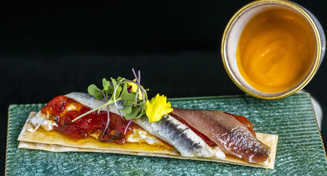 A small tapa of cured ham and fish fillets on a green tile with a glass of amber drink behind