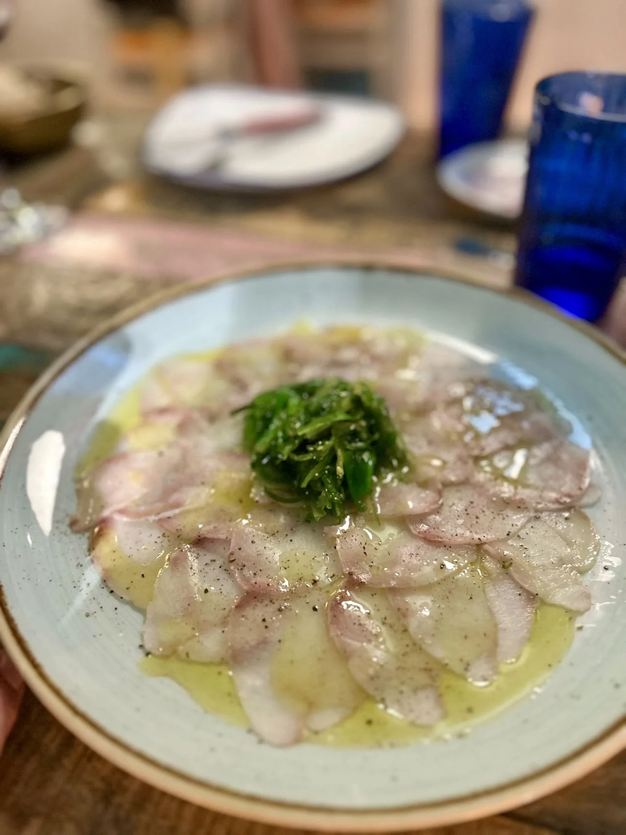 A fish carpaccio drizzled with oil and topped with a sprig of herb