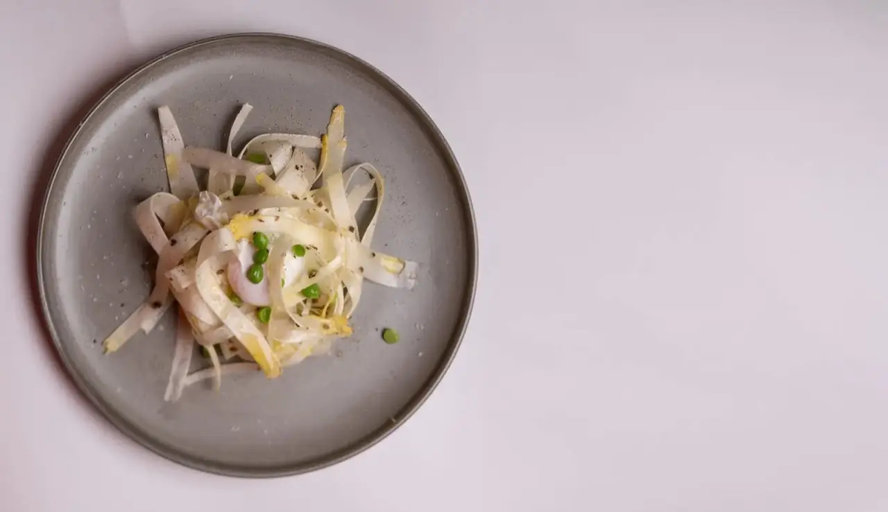 An overhead shot of a delicate dish of shaved white vegetable ribbons with green peas and a poached egg on a grey ceramic plate