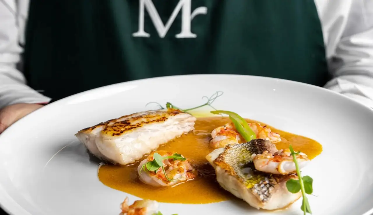A plate of seared white fish and langoustines in an orange bisque-style sauce with pea shoots, held by a server in a green monogrammed apron