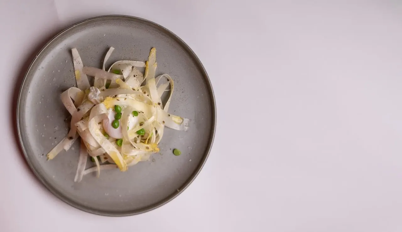 A grey plate of finely sliced pale ingredients with green pieces, seen from above on a pale table