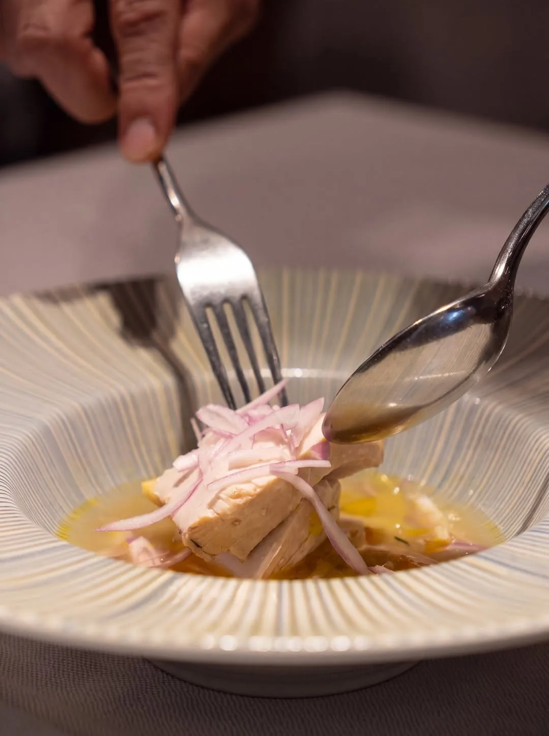 A hand lifting a fork and spoon over a striped white bowl of sliced onion and pale pieces in golden sauce