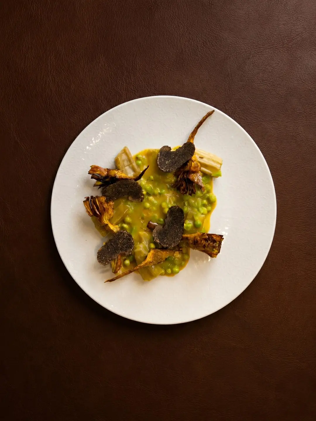Plated dish of green peas and pea puree with roasted baby artichokes and shaved black truffle on a white plate