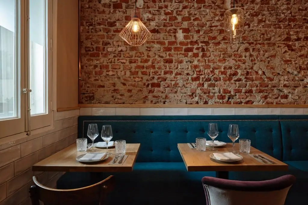Dining room with an exposed red-brick wall, tufted teal banquette, wooden tables set with wine glasses, and copper pendant lights