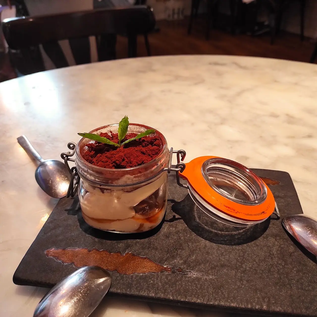 A layered dessert in a glass jar topped with cocoa powder and a mint sprig, served on a slate board with spoons.