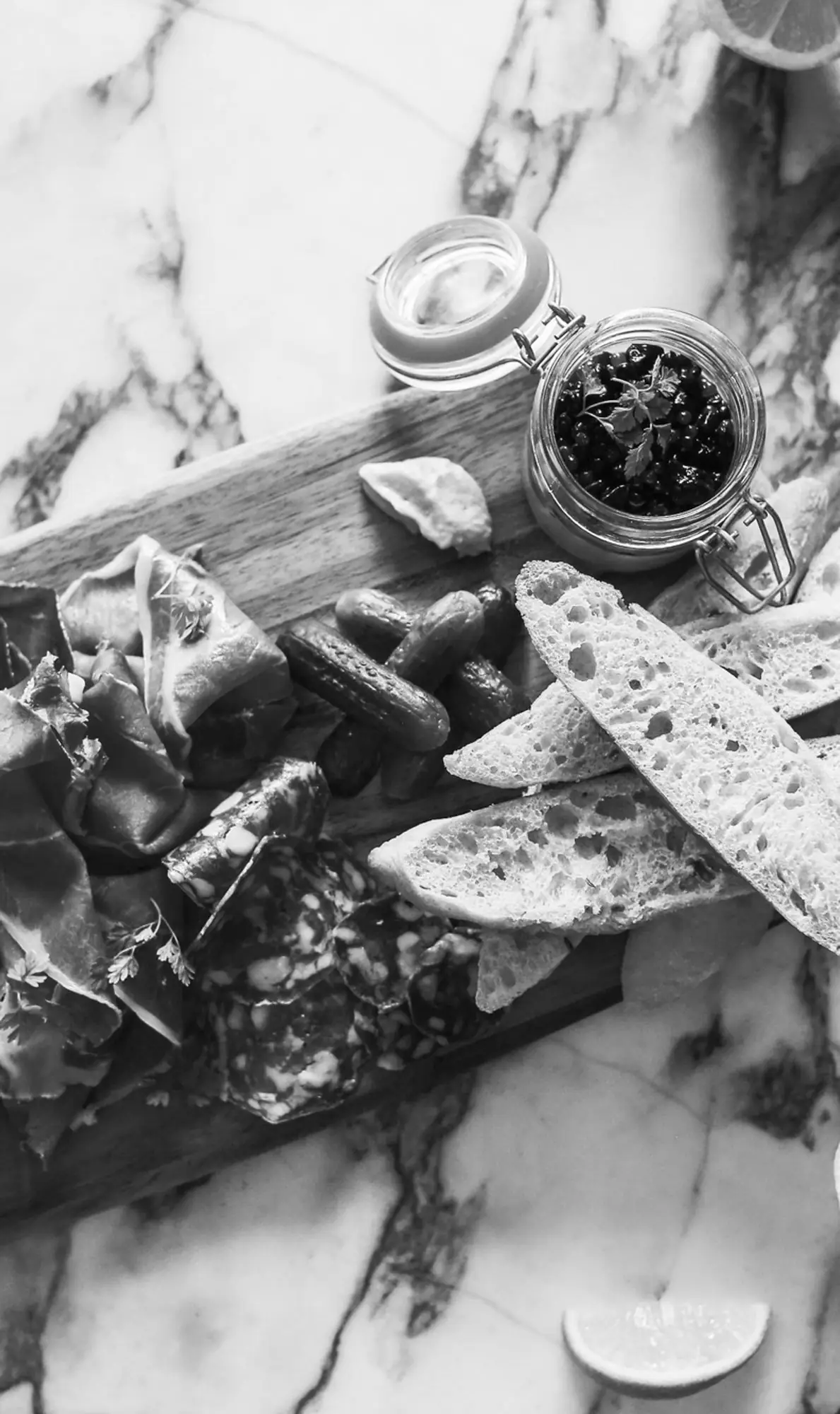 Black-and-white charcuterie board on marble with cured ham, sliced salami, cornichons, toasted bread and a clip-jar of black olives topped with parsley