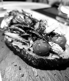 Black-and-white close-up of a toast topped with cherry tomatoes, anchovies and threads of greens on a slate board
