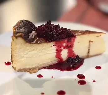 Slice of baked cheesecake on a white plate with red berry compote and berry-syrup droplets