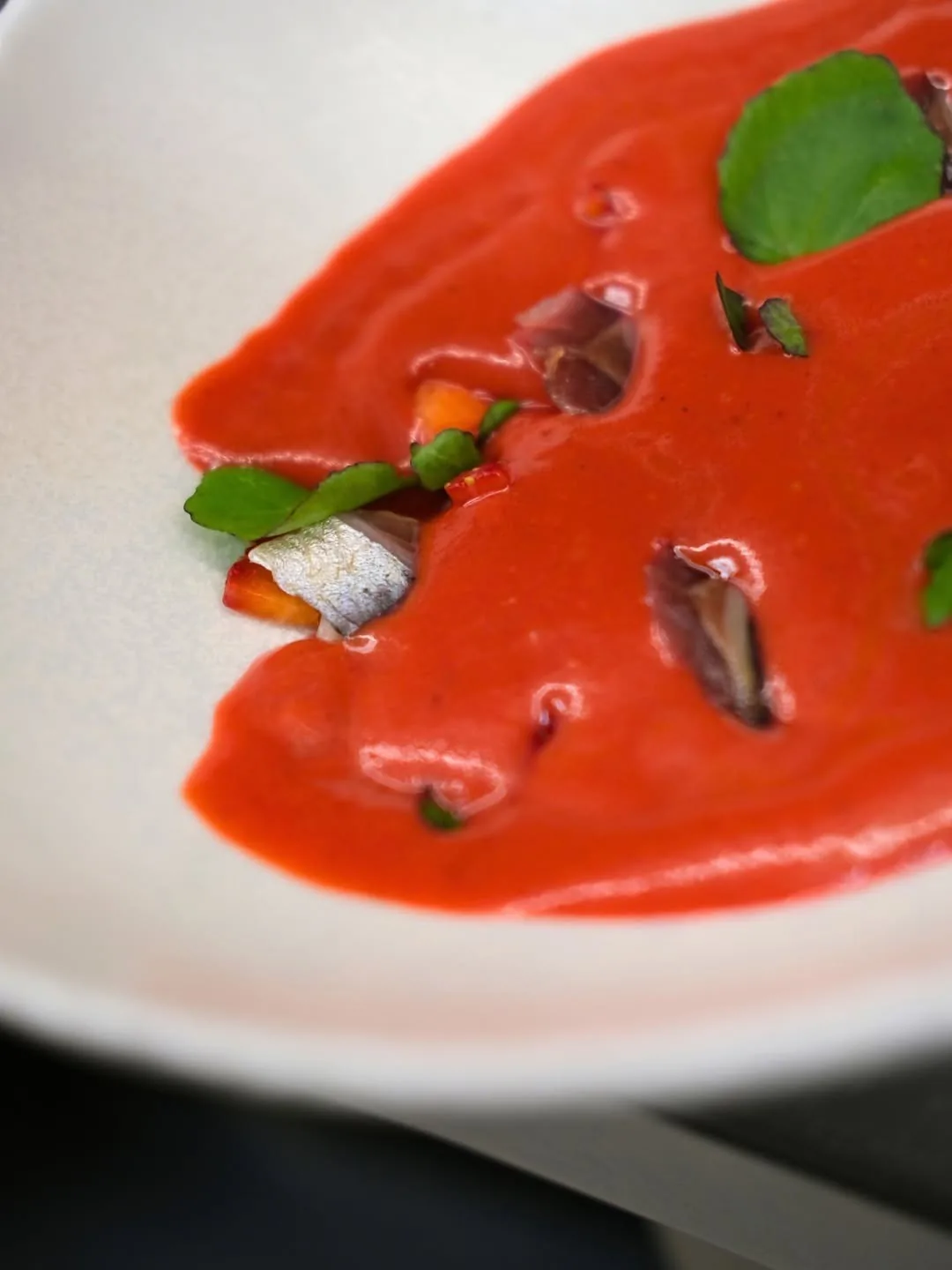 A plate of red soup with small pieces of fish and basil leaves