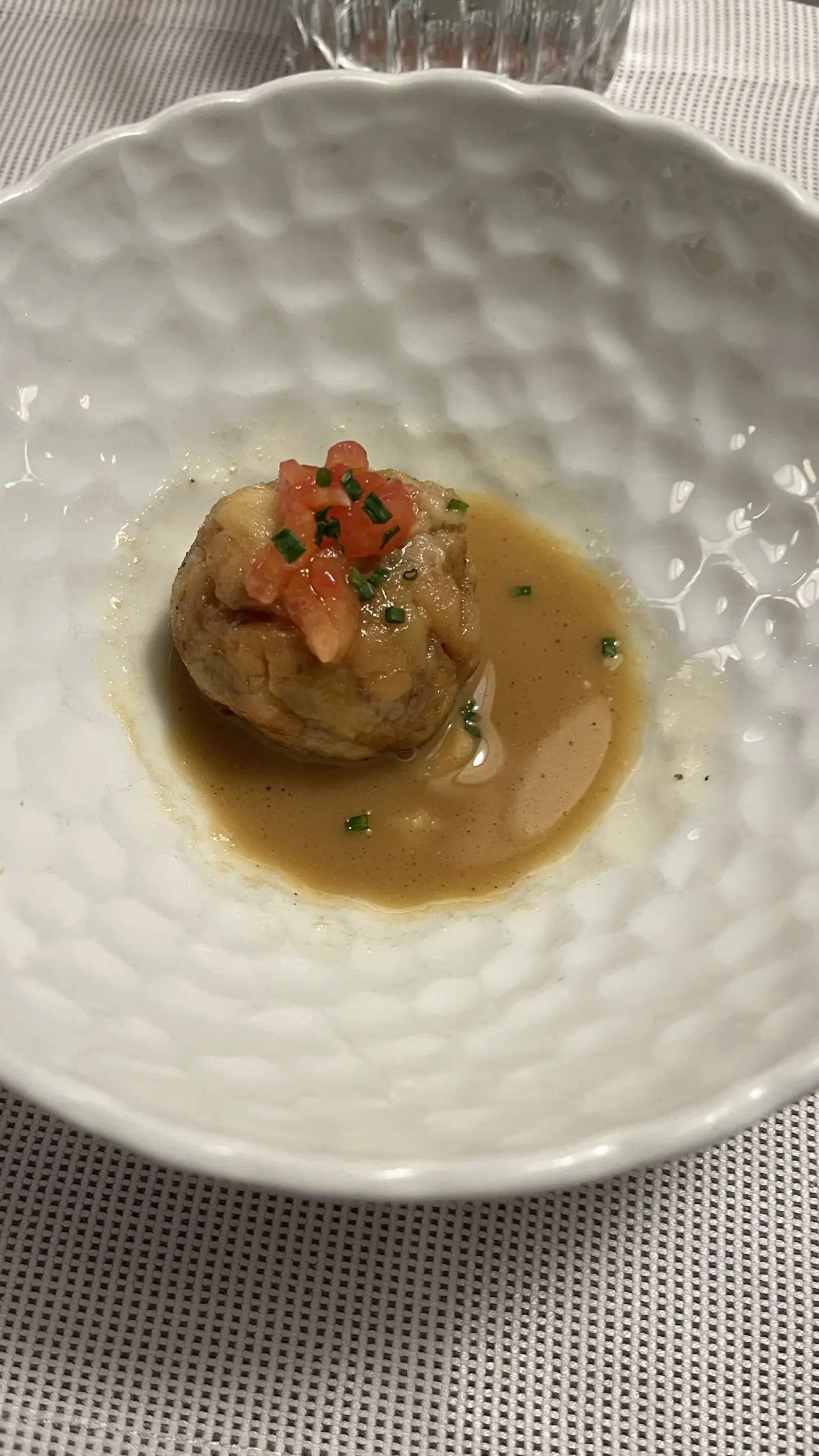 A round croquette-style bite topped with diced tomato and chives in a light brown sauce, in a textured white bowl