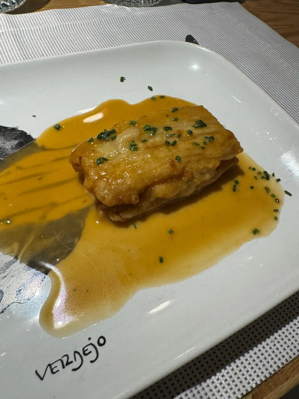A seared fish fillet with chives in a yellow sauce on a plate marked with the restaurant name Verdejo