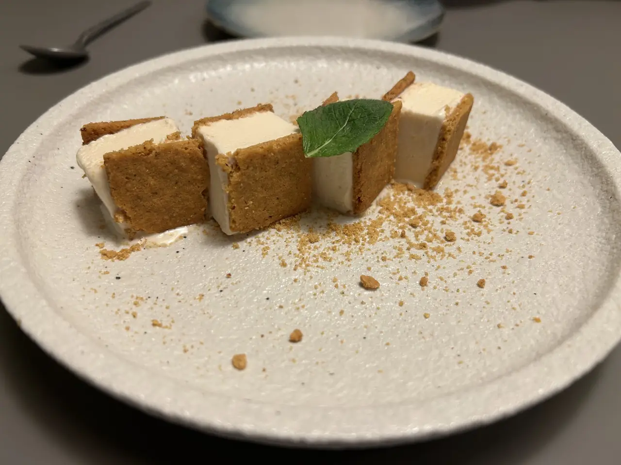 Cubes of layered cream dessert with biscuit crust, crumble, and a mint leaf on a stone plate