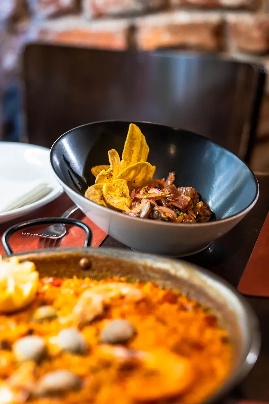 Bowl of ceviche-style beef with fried plantain chips and red onion, with a seafood paella pan in the foreground