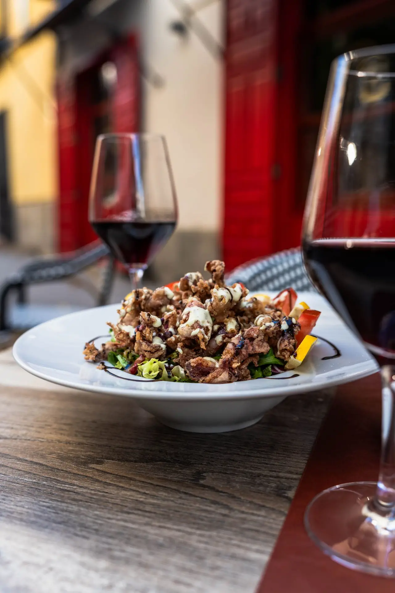 Fried breaded dish with balsamic drizzle on a plate, wine glasses on an outdoor terrace table