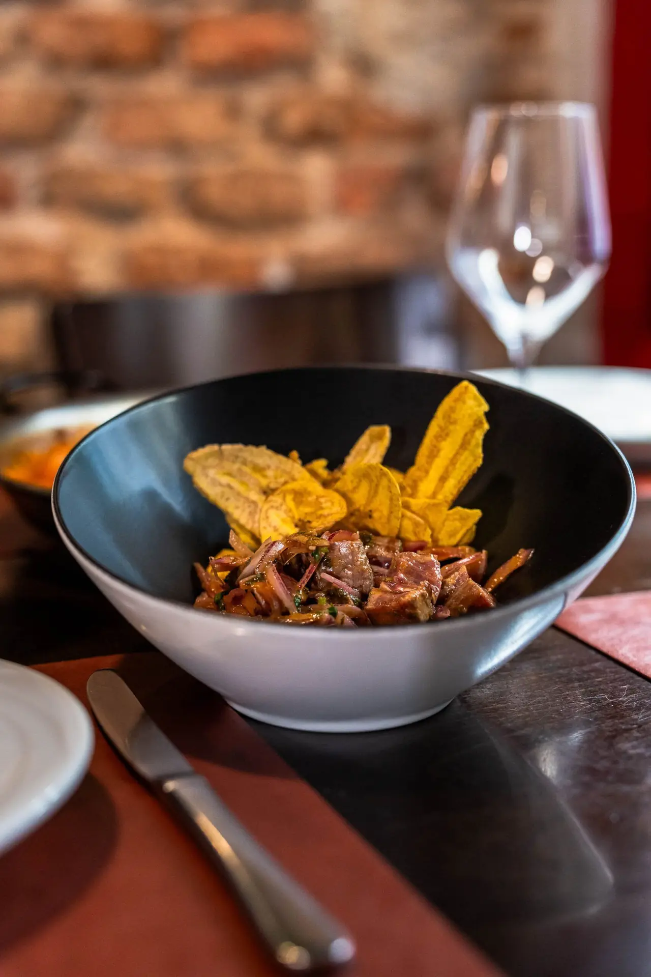 Bowl of marinated beef with fried plantain chips on a restaurant table with wine glass