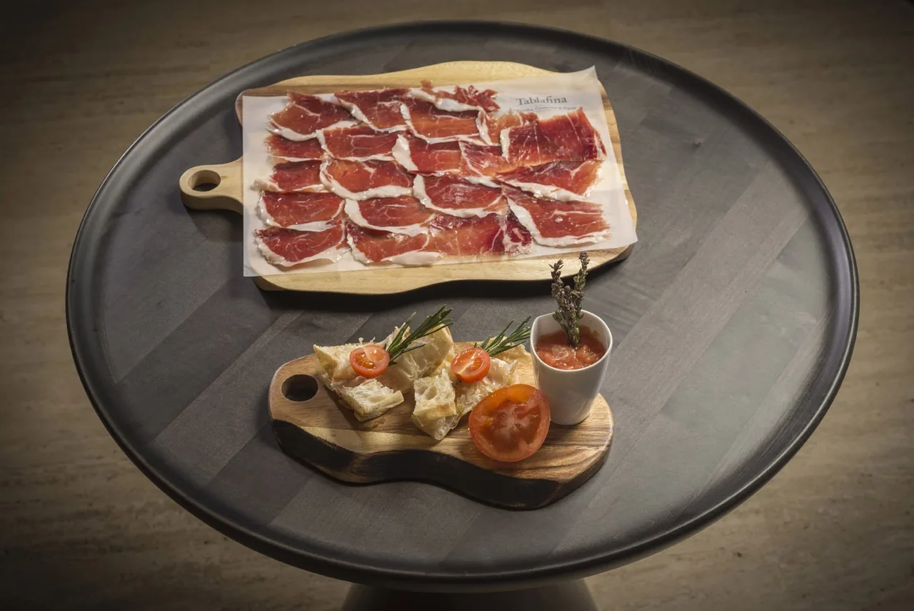 Slices of cured ham on a round dark board beside a wooden board of cheese and small bites