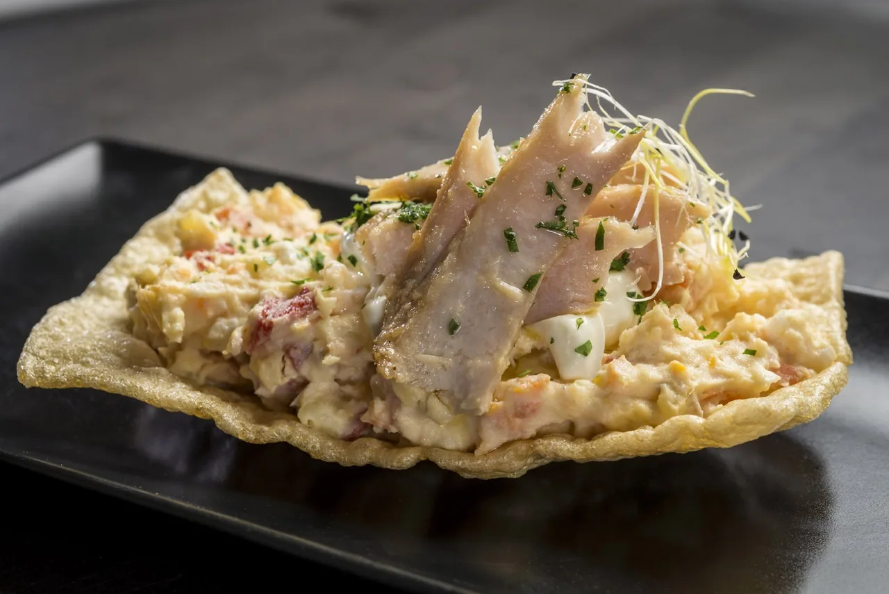 A crisp round base topped with a creamy salad and flaked fish on a dark plate