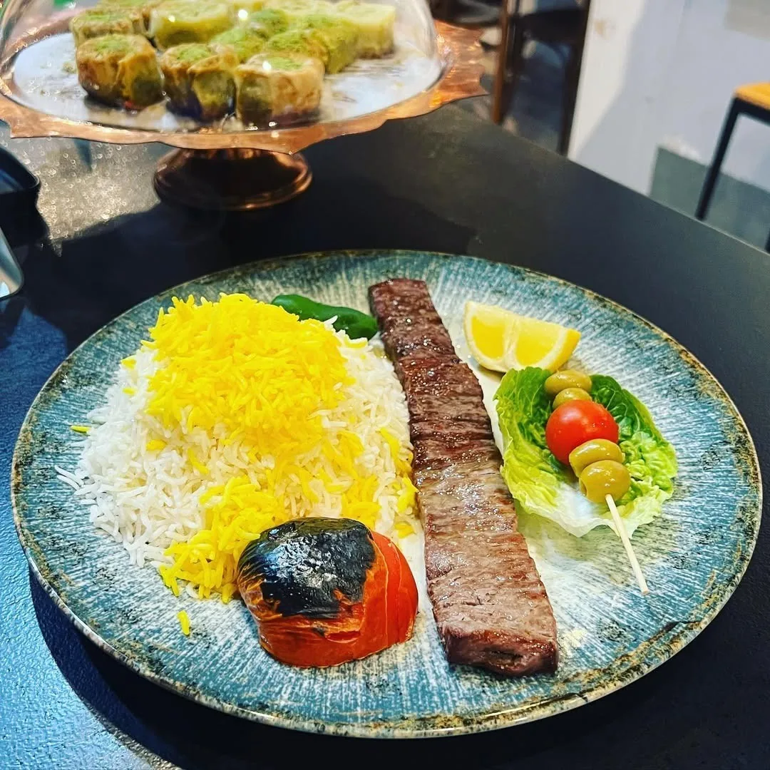 A blue plate with saffron rice, a skewer of minced meat, a grilled tomato and salad leaves
