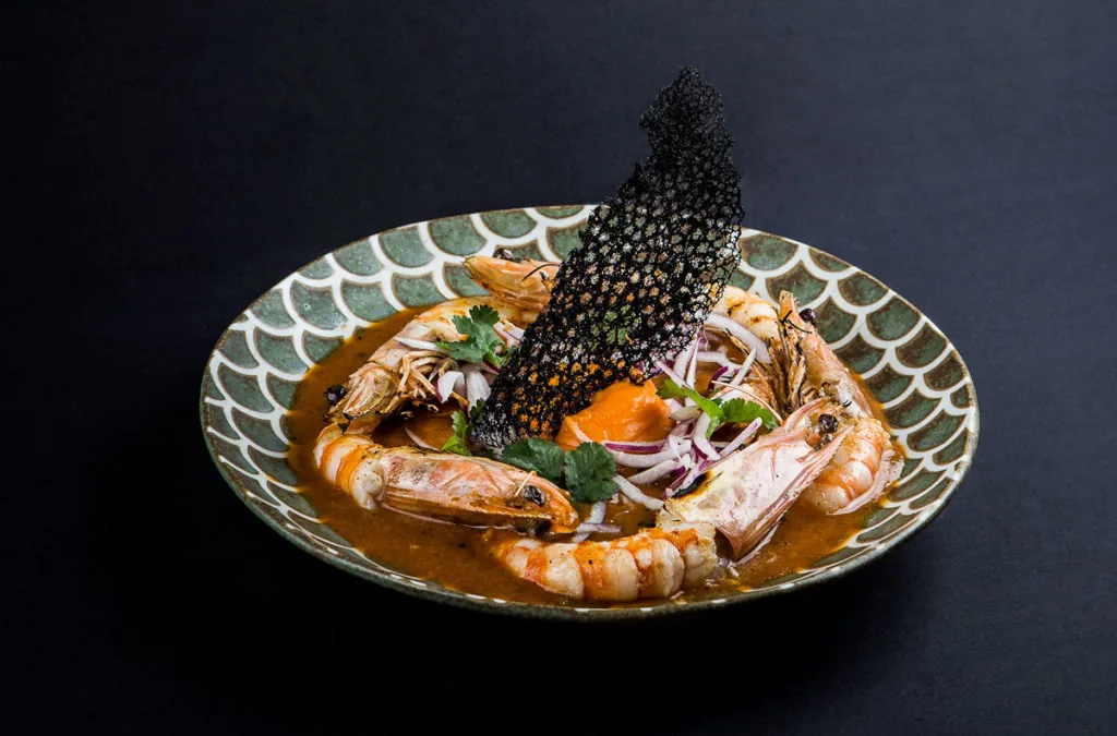 Whole head-on prawns in a rust-orange broth in a green scallop-patterned bowl, topped with a black lattice cracker standing upright, sliced red onion, coriander