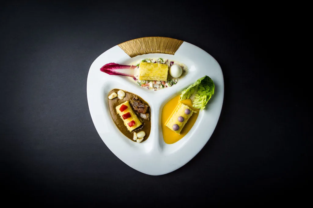 Overhead view of a white three-lobed plate with a gold fan detail, holding three separate layered potato-terrine preparations: one with chopped salad and a whit