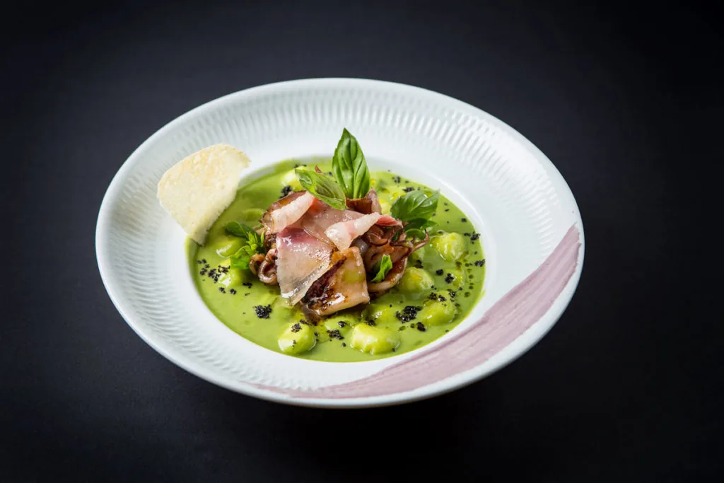Grilled squid and thin slices of cured meat over a bright green pea sauce with gnocchi, black crumb, basil leaves and a pale crisp, in a ribbed white bowl with