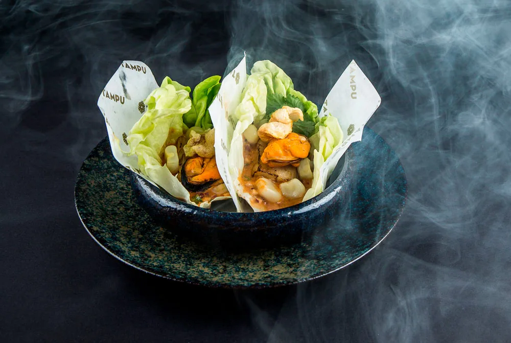 A dark speckled stone bowl steaming heavily, holding lettuce leaves and paper wrappers printed TAMPU filled with mussels, scallops, corn and fried garnish