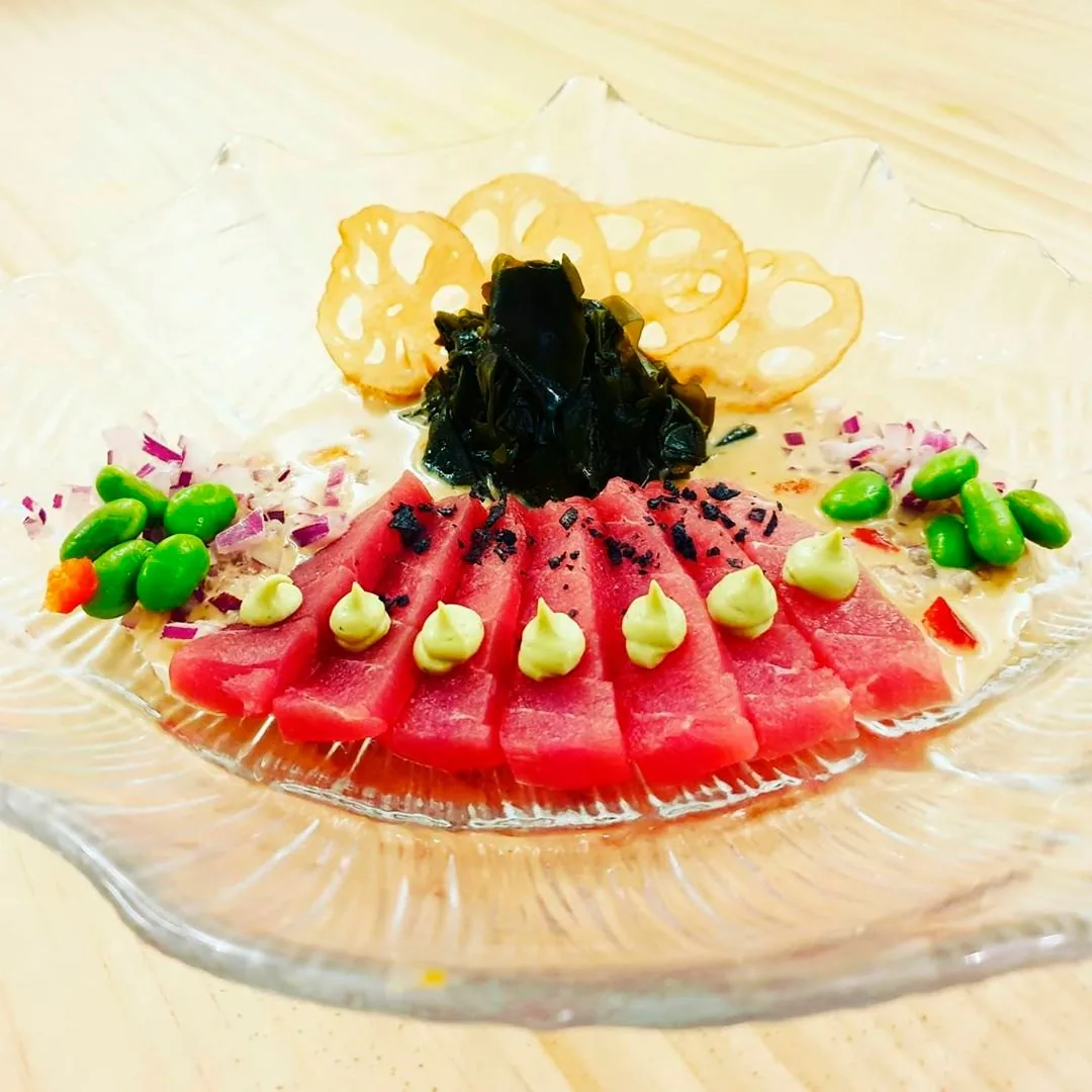 Slices of raw red tuna topped with dark seaweed, green dots and lotus root chips on a glass plate