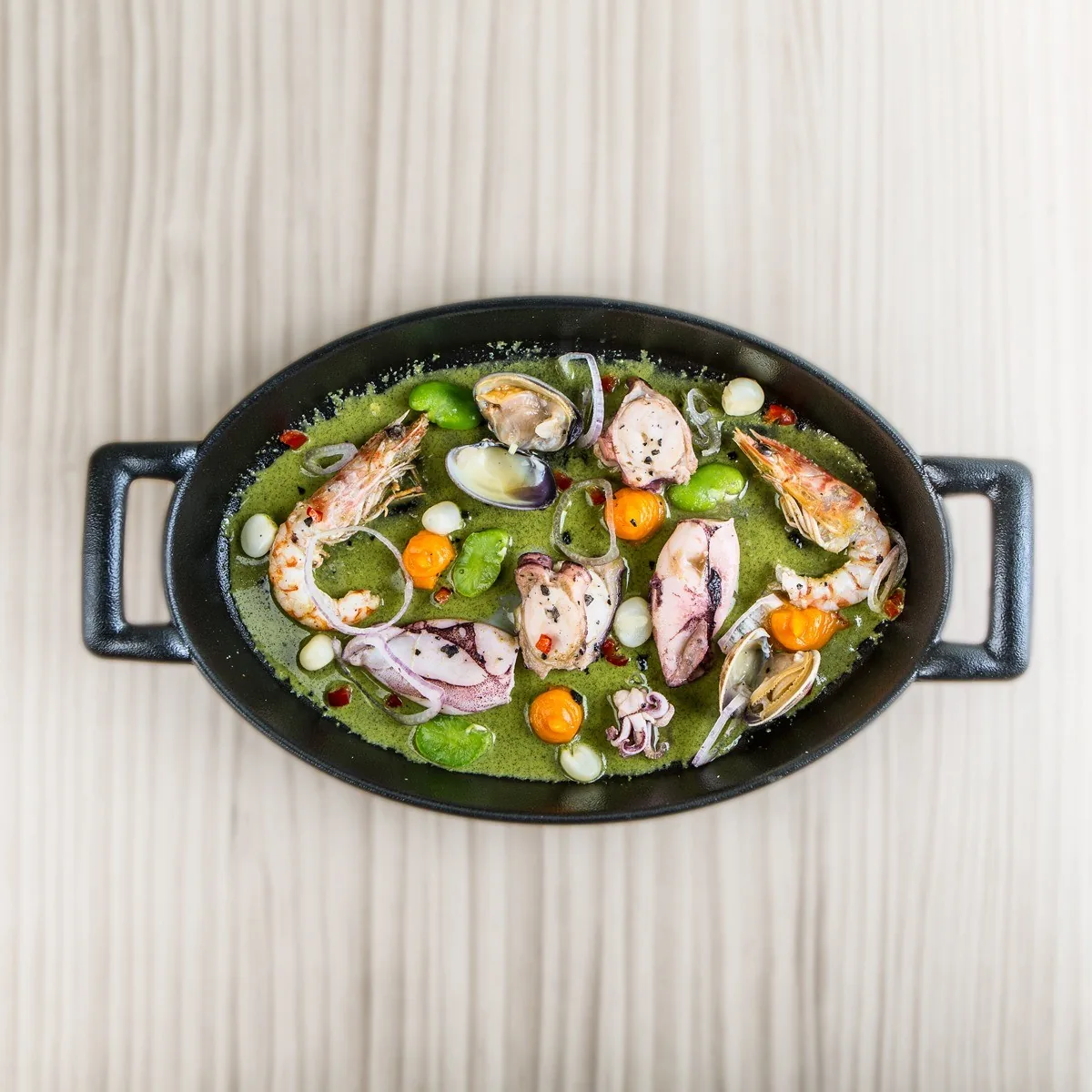 A black pan of seafood and vegetables in a green sauce on a pale surface