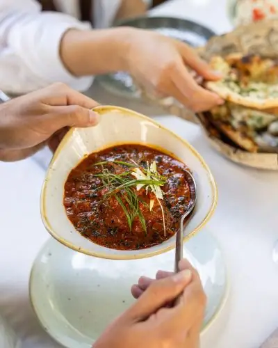 Bowl of dark red curry garnished with slivered ginger and scallion, held over a plate with a basket of naan behind, a spoon dipping in