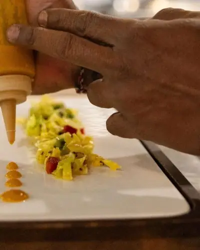 Hands plating a chopped vegetable garnish and drizzling a mustard-colored sauce in dots on a white rectangular plate