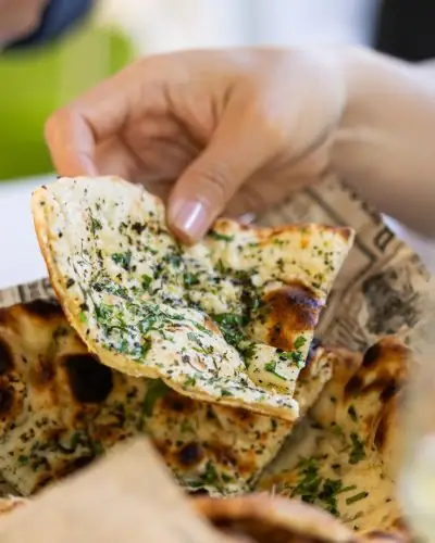 Hand lifting a piece of garlic-and-herb butter naan from a basket of charred flatbread