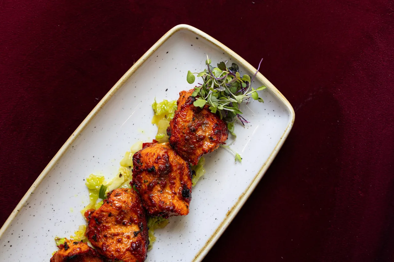 A rectangular plate of glazed grilled chicken pieces garnished with micro herbs on a dark red cloth