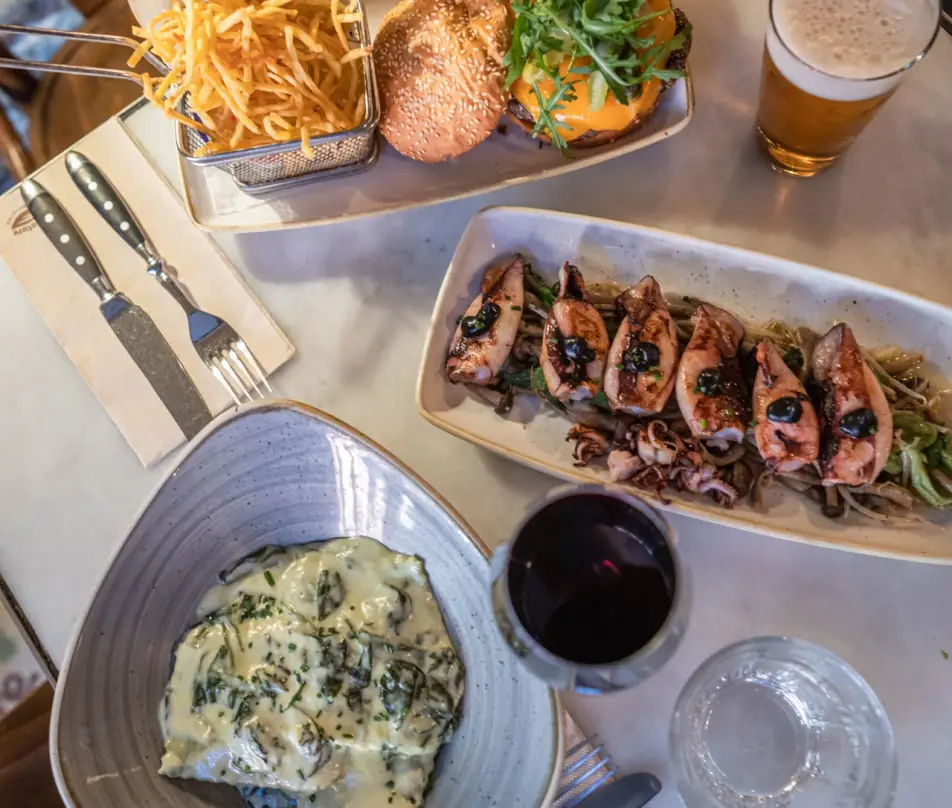 Grilled squid tentacles on a bed of noodles, a burger with fries, and a creamy gratin dish shared on a marble table at Tantarantana